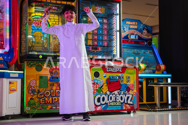 الاستمتاع في العطلة الصيفية، الشعور بالفرح والسعادة، المراكز الترفيهية في المملكة العربية السعودية، ألعاب الأركيد الالكترونية، ولد عربي سعودي خليجي يرتدي الثوب التقليدي ينظر إلى الكاميرا يرفع يديه بإيماءات الفوز والربح