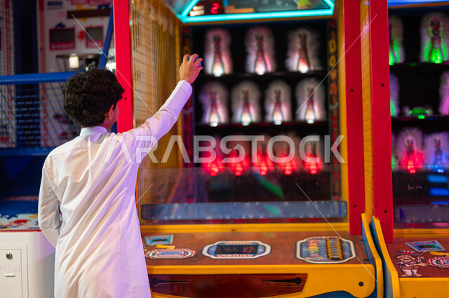 الشعور بالفرح والحماس، الاستمتاع في العطلة الصيفية، المراكز الترفيهية في المملكة العربية السعودية، ألعاب الأركيد الالكترونية، صورة من الخلف لولد عربي سعودي خليجي يرتدي الثوب التقليدي يمسك الكرة يلعب لعبة الرمي والتصويب على الهدف .