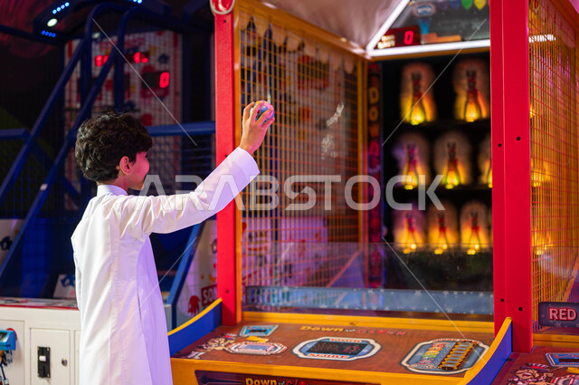 الشاشات الملونة المضيئة، الشعور بالفرح والحماس، الاستمتاع في العطلة الصيفية، المراكز الترفيهية في المملكة العربية السعودية، ألعاب الأركيد الالكترونية، صورة من الجانب لولد عربي سعودي خليجي يرتدي الثوب التقليدي يمسك الكرة يلعب لعبة الرمي والتصويب على الهدف.