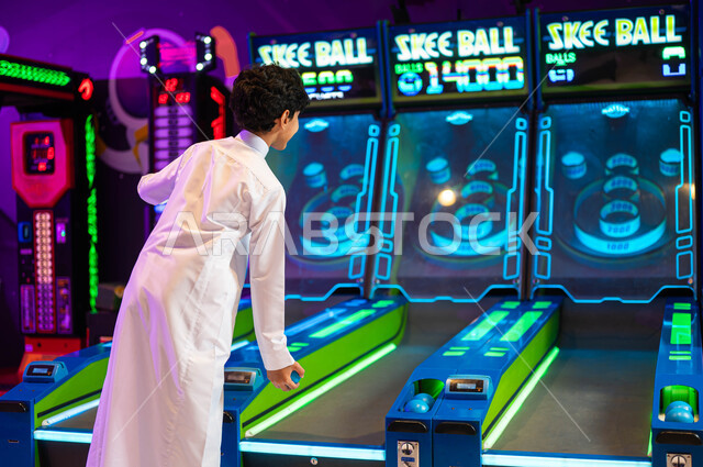 اللعب بألعاب الاركيد الالكترونية المتطورة، لعبة رولينج سكاي بول، المراكز الترفيهية في المملكة، صورة من الخلف لولد عربي سعودي خليجي يرتدي الثوب التقليدي يقضي وقت ممتع في قسم التحديات والمغامرات، قضاء عطلة صيفية متميزة.