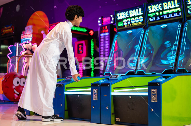 قضاء اوقات ممتعة في قسم التحديات والمغامرات، المراكز الترفيهية في المملكة،  ألعاب الاركيد الالكترونية المتطورة، صورة من الجانب لولد عربي سعودي خليجي يرتدي الثوب التقليدي يقوم باللعب بلعبة رولينج سكاي بول، الاستمتاع في العطلة الصيفية .