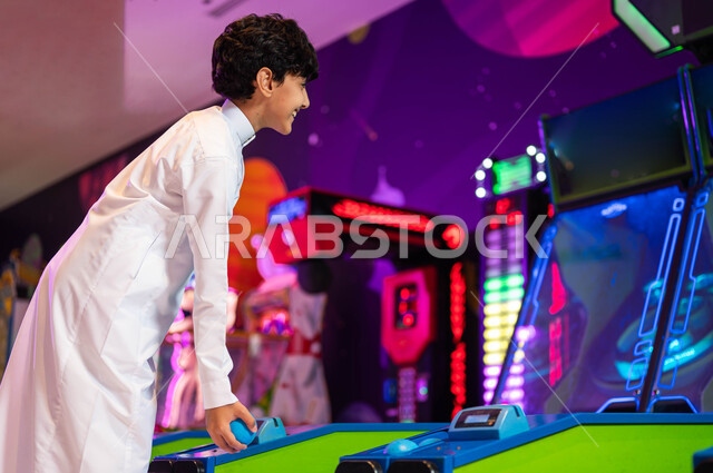 قضاء اوقات ممتعة في قسم التحديات والمغامرات، المراكز الترفيهية في المملكة،  ألعاب الاركيد الالكترونية المتطورة، صورة من الجانب لولد عربي سعودي خليجي يرتدي الثوب التقليدي يقوم باللعب بلعبة رولينج سكاي بول، الاستمتاع في العطلة الصيفية .