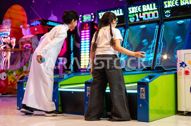 الاستمتاع في اللعب مع الأخوة، التسلية و الترفيه في أرض المرح في العطلة، لعبة رولينج سكاي بول، ألعاب الاركيد الالكترونية المتطورة، صورة من الخلف لولد عربي سعودي خليجي مع أخته يقضيان وقت ممتع في مدينة الملاهي، قسم التحديات والمغامرات.