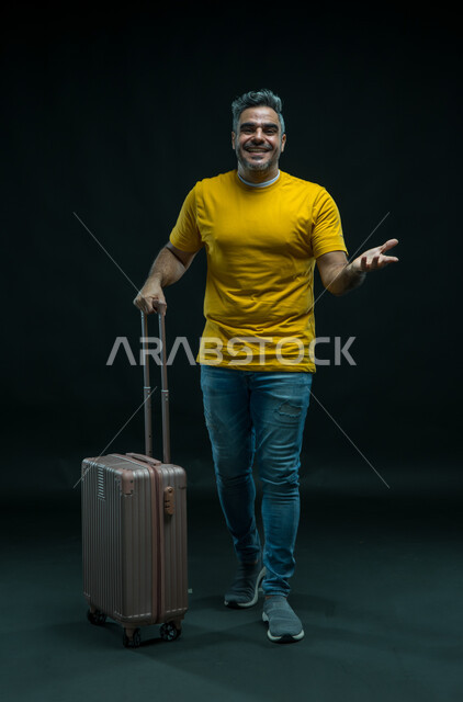 الاستمتاع في إجازة الصيف, السؤال عن شيء ما بإيماءات السرور، الاستعداد لقضاء رحلة صيفية ممتعة, بورتريه لرجل عربي سعودي خليجي مبتسم يرتدي الزي الكاجوال يمسك حقيبة السفر, النظر إلى الكاميرا والشعور بالحماس والفرح، صورة الجسم بالطول الكامل, خلفية سوداء.