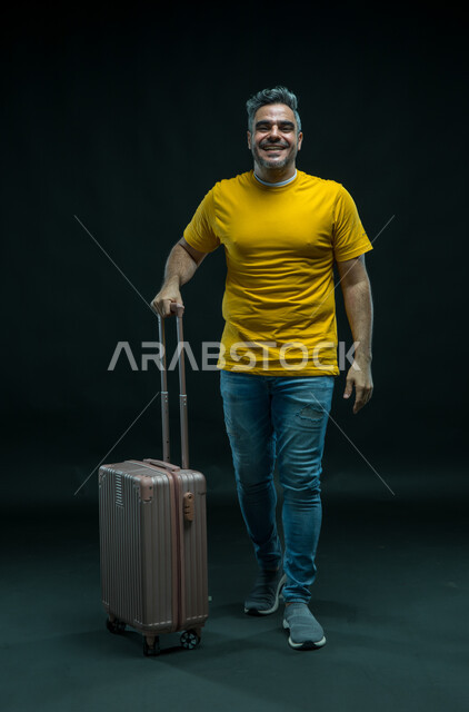 السياحة في إجازة الصيف, الاستعداد لقضاء رحلة صيفية ممتعة, إيماءات السرور والسعادة, بورتريه لرجل عربي سعودي خليجي مبتسم يرتدي الزي الكاجوال ينظر إلى الكاميرا يمسك حقيبة السفر, الشعور بالحماس والفرح، صورة الجسم بالطول الكامل, خلفية سوداء.