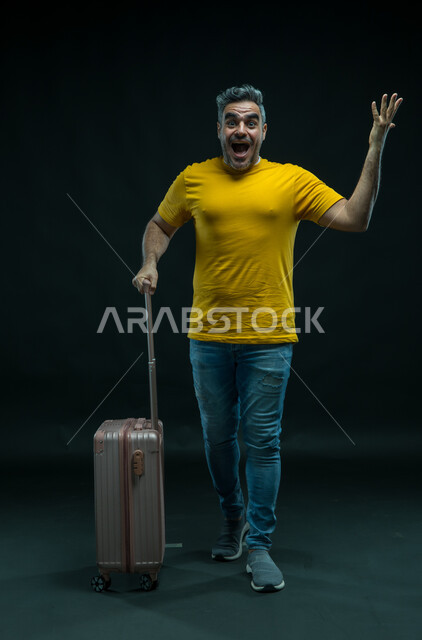 إيماءات الاندهاش والذهول, السياحة في إجازة الصيف,  بورتريه لرجل عربي سعودي خليجي يرتدي الزي الكاجوال يمسك حقيبة السفر, الاستعداد لقضاء رحلة صيفية ممتعة, النظر إلى الكاميرا والشعور بالحماس والفرح، صورة الجسم بالطول الكامل,  خلفية سوداء.
