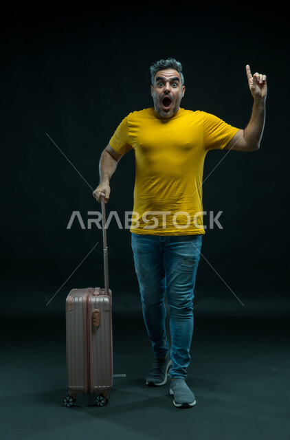 النظر إلى الكاميرا بإيماءات الذهول والإشارة إلى شيء ما, السياحة في إجازة الصيف,  بورتريه لرجل عربي سعودي خليجي يرتدي الزي الكاجوال يمسك حقيبة السفر, الاستعداد لقضاء رحلة صيفية ممتعة, الشعور بالحماس والفرح، صورة الجسم بالطول الكامل,  خلفية سوداء.