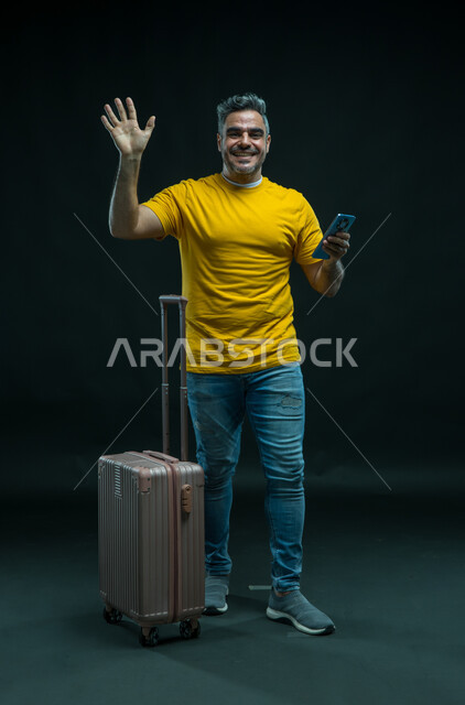 إيماءات الشكر وعدم الحاجه للمساعده، التلويح باليد للتعبير عن الوداع والانطلاق لبدء الرحلة، بورتريه لرجل عربي خليجي سعودي مبتسم يرتدي الزي الكاجوال ينظر إلى الكاميرا يقف بجانب حقيبة السفر يستخدم الهاتف المحمول, صورة الجسم بالطول الكامل ، خلفية سوداء