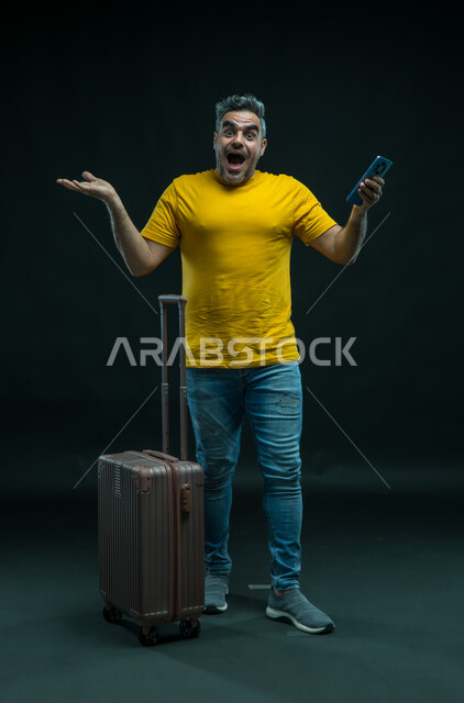 التعجب من الخدمات المتطورة عبر الهاتف المحمول، النظر إلى الكاميرا بإيماءات الاندهاش والذهول، الاستعداد لقضاء رحلة صيفية ممتعة, بورتريه لرجل عربي سعودي خليجي يرتدي الزي الكاجوال يقف أمام حقيبة السفر يمسك الجوال, صورة الجسم بالطول الكامل, خلفية سوداء.