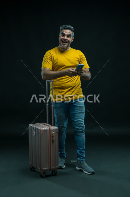 الدفع و تأكيد الحجوزات من خلال تطبيقات الهاتف الذكي، النظر إلى الكاميرا بإيماءات الاندهاش، الاستعداد لقضاء رحلة صيفية ممتعة, بورتريه لرجل عربي سعودي خليجي يرتدي الزي الكاجوال يمسك حقيبة السفر يستخدم الهاتف المحمول, صورة الجسم بالطول الكامل, خلفية سوداء.