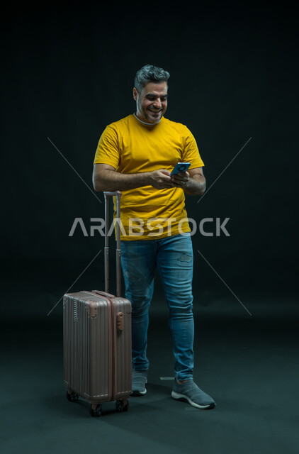 الحجز الفندقي عبر الجوال، استخدام التقنيات الحديثة، الاستعداد لقضاء رحلة صيفية ممتعة, بورتريه لرجل عربي سعودي خليجي مبتسم يرتدي الزي الكاجوال يقف امام حقيبة السفر يمسك الهاتف المحمول, صورة الجسم بالطول الكامل, خلفية سوداء.