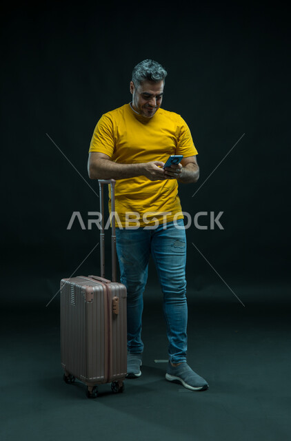 استخدام التقنيات الحديثة، الحجز الفندقي عبر الجوال، الاستعداد لقضاء رحلة صيفية ممتعة, بورتريه لرجل عربي سعودي خليجي مبتسم يرتدي الزي الكاجوال يقف أمام حقيبة السفر يمسك الهاتف المحمول, صورة الجسم بالطول الكامل, خلفية سوداء.