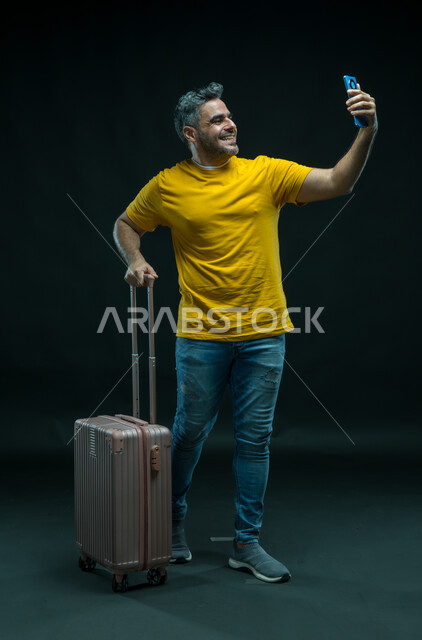 التقاط صور السيلفي التذكارية، توثيق اللحظات السعيدة في إجازة الصيف, الاستعداد لقضاء رحلة صيفية ممتعة, بورتريه لرجل عربي سعودي خليجي مبتسم يرتدي الزي الكاجوال يمسك حقيبة السفر يستخدم الهاتف المحمول, صورة الجسم بالطول الكامل, خلفية سوداء.
