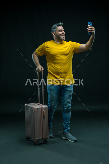 توثيق اللحظات السعيدة في إجازة الصيف, الاستعداد لقضاء رحلة صيفية ممتعة, بورتريه لرجل عربي سعودي خليجي مبتسم يرتدي الزي الكاجوال يمسك حقيبة السفر يستخدم الهاتف المحمول, التقاط صور السيلفي التذكارية، صورة الجسم بالطول الكامل, خلفية سوداء.
