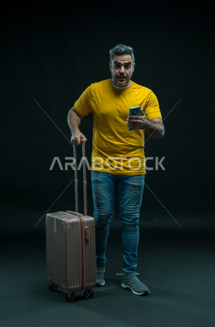 الانزعاج لحدوث شيء ما, النظر إلى الكاميرا بإيماءات الاستغراب، الاستعداد لقضاء رحلة صيفية ممتعة, بورتريه لرجل عربي سعودي خليجي يرتدي الزي الكاجوال يمسك حقيبة السفريستخدم الهاتف المحمول, صورة الجسم بالطول الكامل, خلفية سوداء.