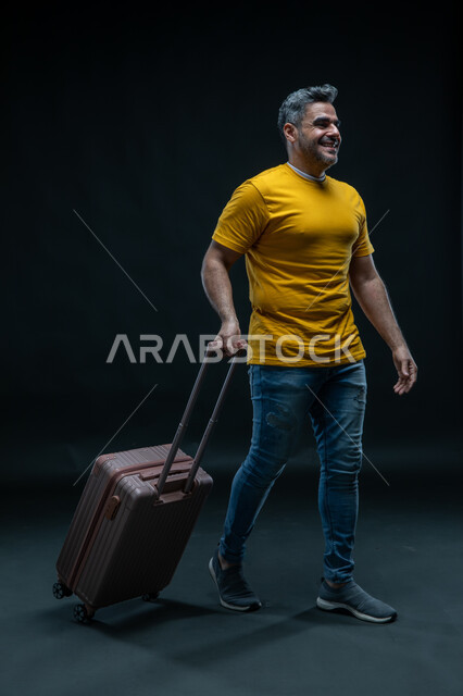 الاستعداد للانطلاق وبدء الرحلة، الاستمتاع في قضاء إجازة الصيف، بورتريه لرجل عربي خليجي سعودي مبتسم يرتدي الزي الكاجوال يمسك حقيبة السفر, الحماس للذهاب في جولة سياحية، صورة الجسم بالطول الكامل ، خلفية سوداء