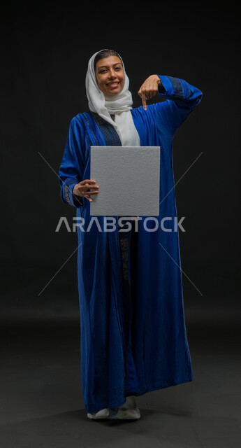لوحة بيضاء مربعة فارغة، استخدام لوحات توضيحية تعريفية، إعلانات ودعايات وعروض تسويقية، بورتريه لإمرأة عربية خليجية سعودية مبتسمة ترتدي حجاب وعباءة زرقاء تحمل بيدها موك اب مربع أبيض تشير إليه بإصبع السبابة تنظر الى الكاميرا، صورة الجسم بالكامل، خلفية سوداء