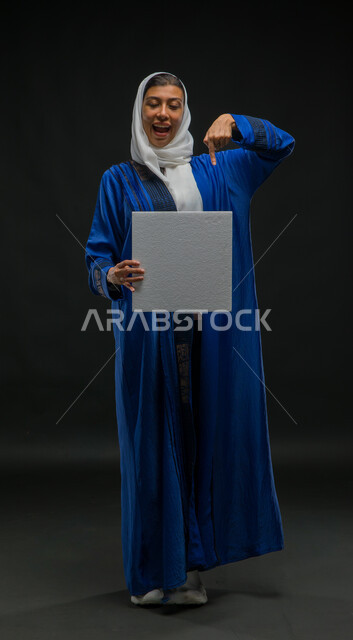 تعابير الحماس لعرض إعلانات ودعايات وعروض تسويقية، بورتريه لإمرأة عربية خليجية سعودية متحمسة ترتدي حجاب وعباءة زرقاء تحمل بيدها موك اب مربع أبيض تشير إليه بإصبع السبابة، استخدام لوحات توضيحية تعريفية، لوحة بيضاء مربعة فارغة، صورة الجسم بالكامل، خلفية سوداء