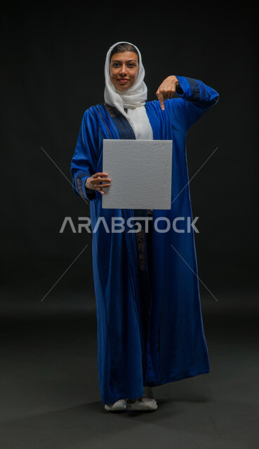 استخدام لوحات توضيحية تعريفية، لوحة بيضاء مربعة فارغة، إعلانات ودعايات وعروض تسويقية، بورتريه لإمرأة عربية خليجية سعودية مبتسمة ترتدي حجاب وعباءة زرقاء تحمل بيدها موك اب مربع أبيض تشير إليه بإصبع السبابة تنظر الى الكاميرا، صورة الجسم بالكامل، خلفية سوداء