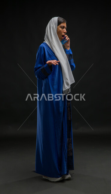 السؤال والاستفسار عن أمر ما بإنفعالات وتعابير الغضب والقلق, الشعور بالانزعاج لسماع الأخبار السيئة, بورتريه لامرأة عربية خليجية سعودية محجبة ترتدي عباءة زرقاء تقوم بإجراء مكالمة هاتفية بإسخدام الجوال، صورة الجسم بالطول الكامل، خلفية سوداء