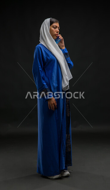 تعابير التركيز والاندماج بالتحدث عبر الهاتف المحمول، قضاء اوقات ممتعة بتبادل الاخبار والأحاديث، بورتريه لامرأة عربية خليجية سعودية محجبة ترتدي عباءة زرقاء تقوم بإجرء مكالمة هاتفية، محادثات ودردشات مع الاصدقاء، صورة الجسم بالكامل، خلفية سوداء