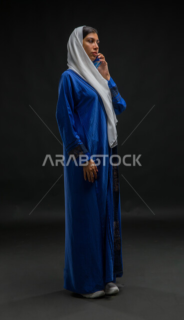 إيماءات الاندماج والتركيز بالتحدث عبر الهاتف المحمول، قضاء اوقات ممتعة بتبادل الاخبار والأحاديث، بورتريه لامرأة عربية خليجية سعودية محجبة ترتدي عباءة زرقاء تقوم بإجرء مكالمة هاتفية، محادثات ودردشات مع الاصدقاء، صورة الجسم بالكامل، خلفية سوداء