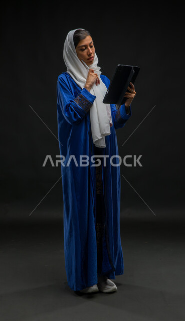 التسوق الإلكتروني الآمن، تعابير وإيماءات التفكير والحيرة, مفهوم إدارة المشاريع عن بعد, بورتريه لإمرأة عربية خليجية سعودية محجبة ترتدي عباءة زرقاء تقوم بانجاز مهام العمل عبر جهاز التابلت, إستخدام اجهزة تقنية حديثة، صورة الجسم بالكامل، خلفية سوداء