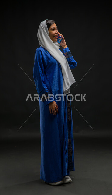 Facial gestures indicating happiness and pleasure in making a phone call, portrait of a veiled Saudi Arabian Gulf woman wearing a blue abaya talking on a mobile phone, spending enjoyable times exchanging news and conversations, conversations and chats with friends, full body image, black background