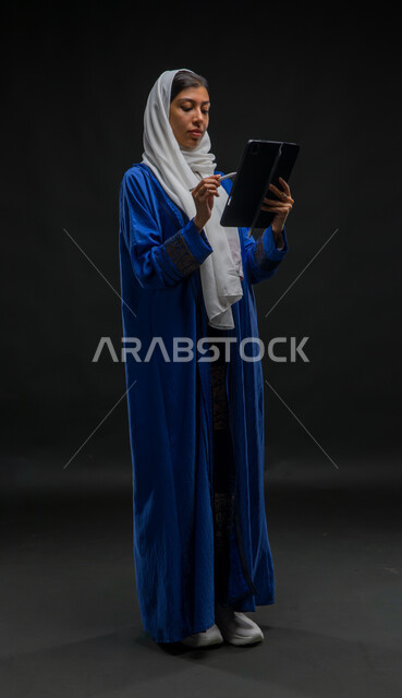 إيماءات وتعابير التركيز والاندماج, إستخدام اجهزة تقنية حديثة، بورتريه لإمرأة عربية خليجية سعودية محجبة ترتدي عباءة زرقاء تقوم بانجاز مهام العمل عبر جهاز التابلت, مفهوم إدارة المشاريع عن بعد, التسوق الإلكتروني الآمن، صورة الجسم بالكامل، خلفية سوداء