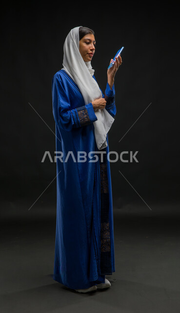 تسجيل مقطع صوتي بإستخدام الهاتف المحمول، الدردشة والتحدث مع الاقارب، بورتريه لإمرأة عربية خليجية سعودية محجبة ترتدي عباءة زرقاء تقوم بالاستماع الى المقاطع الصوتية عبر الجوال، قضاء وقت الفراغ بالتواصل مع الصديقات، صورة الجسم بالكامل، خلفية سوداء
