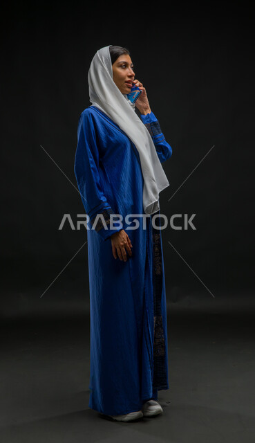 قضاء اوقات ممتعة بتبادل الاخبار والأحاديث، ايماءات وجه تدل على السعادة و السرور بإجراء مكالمة هاتفية، بورتريه لامرأة عربية خليجية سعودية محجبة ترتدي عباءة زرقاء تقوم بالتحدث عبر الهاتف المحمول، محادثات ودردشات مع الاصدقاء، صورة الجسم بالكامل، خلفية سوداء