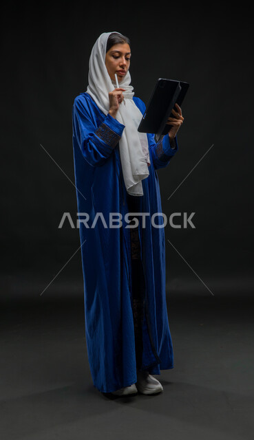 مفهوم إدارة المشاريع عن بعد, بورتريه لإمرأة عربية خليجية سعودية محجبة ترتدي عباءة زرقاء تقوم بانجاز مهام العمل عبر جهاز التابلت, تعابير وإيماءات التفكير والحيرة, إستخدام اجهزة تقنية حديثة، التسوق الإلكتروني الآمن، صورة الجسم بالكامل، خلفية سوداء