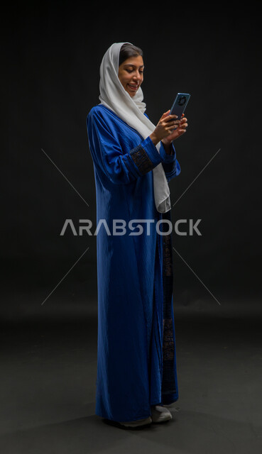الدردشة والتواصل مع التواصل مع الأصدقاء اونلاين، متابعة أخبار وسائل التواصل الاجتماعي باستخدام الجوال، بورتريه لإمرأة عربية خليجية سعودية مبتسمة ترتدي حجاب وعباءة زرقاء تمسك بيديها الهاتف المحمول، تعابير وايماءات الفرح والسرور، صورة الجسم بالكامل، خلفية سوداء