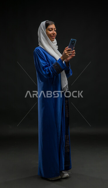 تصفح وسائل التواصل الاجتماعي باستخدام الجوال، بورتريه لإمرأة عربية خليجية سعودية مبتسمة ترتدي حجاب وعباءة زرقاء تمسك بيديها الهاتف المحمول، تعابير وايماءات الفرح والسرور، الدردشة والتواصل مع التواصل مع الأصدقاء اونلاين، صورة الجسم بالكامل، خلفية سوداء