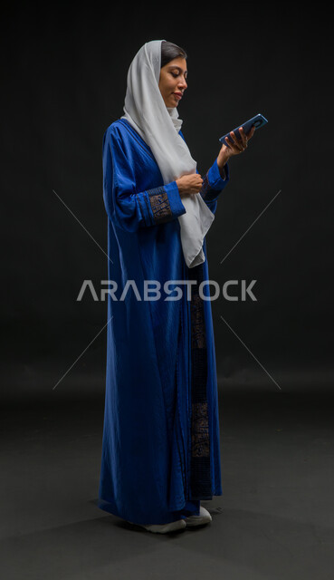 إرسال الرسائل النصية والصوتية عبر الجوال، الدردشة والتحدث مع الاقارب، بورتريه لإمرأة عربية خليجية سعودية محجبة ترتدي عباءة زرقاء تقوم بتسجيل مقطع صوتي بإستخدام الهاتف المحمول،  قضاء وقت الفراغ بالتواصل مع الصديقات، صورة الجسم بالكامل، خلفية سوداء