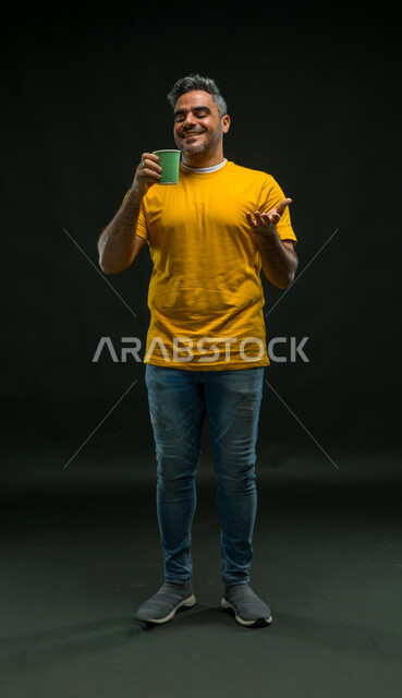 الاستمتاع بمذاق القهوة الزاكي، الحيوية والنشاط عند شرب الكافيين، تناول المشروبات الساخنة، بورتريه لرجل عربي سعودي خليجي مبتسم يرتدي الزي الكاجوال يحمل بيده كوب ورقي، الشعور بالانتعاش والفرح، صورة الجسم بالطول الكامل، خلفية سوداء