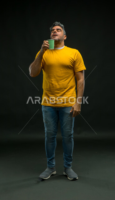 إيماءات الاستمتاع بتناول المشروبات الساخنة، استنشاق رائحة القهوة المنعشة، الحيوية والنشاط عند شرب الكافيين، بورتريه لرجل عربي سعودي خليجي مبتسم يرتدي الزي الكاجوال يحمل بيده كوب ورقي، الشعور بالانتعاش والفرح، صورة الجسم بالطول الكامل، خلفية سوداء .