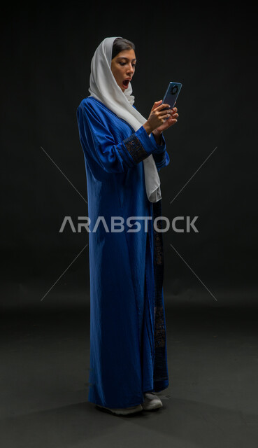 The image of the body is full, the feeling of disturbing to see the sudden and shocking news via mobile, portrait for a veiled Arab Gulf woman who wears a blue cloak that uses a mobile phone, expressions of surprise and amazement, black background