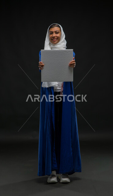 عرض لوحة بيضاء مربعه فارغة بإيماءات البشاشة والسرور، العروض والإعلانات التسويقية, بورتريه لسيدة عربية خليجية سعودية مبتسمة ترتدي عباءة زرقاء تمسك بيديها موك اب مربع أبيض, لوحات توضيحية ووسائل تعليمية, صورة الجسم بالطول الكامل, خلفية سوداء