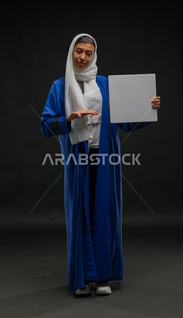 لوحات توضيحية ووسائل تعليمية, الإشارة إلى لوحة بيضاء مربعه فارغة، العروض والإعلانات التسويقية, بورتريه لسيدة عربية خليجية سعودية ترتدي عباءة زرقاء تمسك بيديها موك اب مربع أبيض, صورة الجسم بالطول الكامل, خلفية سوداء