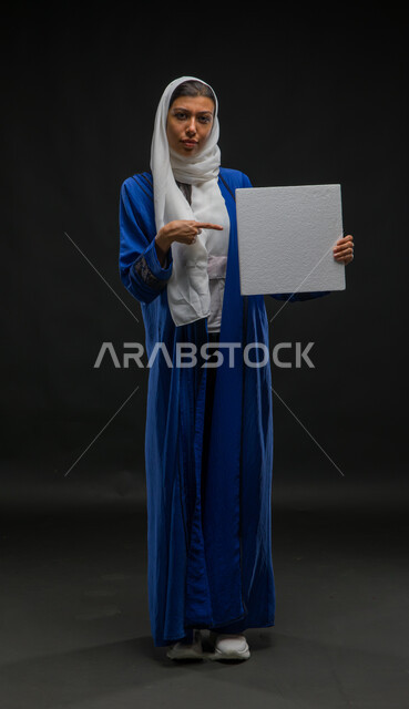 الإشارة إلى موك أب مربع أبيض, إعلانات وعروض تسويقية, بورتريه لإمرأة عربية خليجية سعودية مبتسمة ترتدي عباءة زرقاء تمسك بيدها لوحة بيضاء مربعة فارغة وتنظر إلى الكاميرا, لوحات توضيحية ووسائل تعليمية, صورة الجسم بالطول الكامل, خلفية سوداء