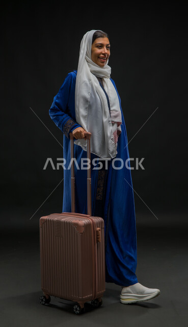 الاستمتاع بالرحلات السياحية في وقت العطلة الصيفية, بورتريه لإمرأة عربية خليجية سعودية مبتسمة ترتدي عباءة زرقاء تمسك بيدها حقيبة السفر, الاستعداد للانطلاق وبدء الرحلة بإيماءات السرور, صورة الجسم بالطول الكامل, خلفية سوداء