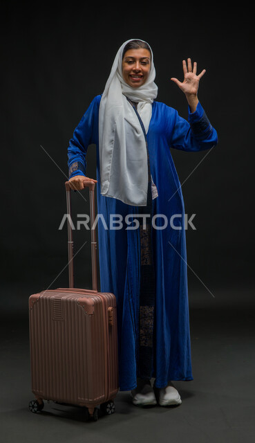 قضاء وقت ممتع مع الأصدقاء, التلويح باليد والتعبير عن الوداع, ايماءات السرور والسعادة بالرحلة السياحية في وقت الإجازة, بورتريه لإمرأة عربية خليجية سعودية مبتسمة ترتدي عباءة زرقاء تمسك بيدها حقيبة السفر وتنظر إلى الكاميرا, صورة لكامل الجسم, خلفية سوداء