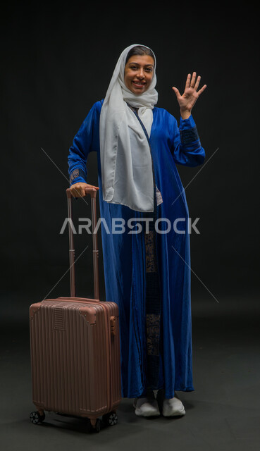 قضاء وقت ممتع مع الأصدقاء, التلويح باليد والتعبير عن الوداع, ايماءات السرور والسعادة بالرحلة السياحية في وقت الإجازة, بورتريه لإمرأة عربية خليجية سعودية مبتسمة ترتدي عباءة زرقاء تمسك بيدها حقيبة السفر وتنظر إلى الكاميرا, صورة لكامل الجسم, خلفية سوداء