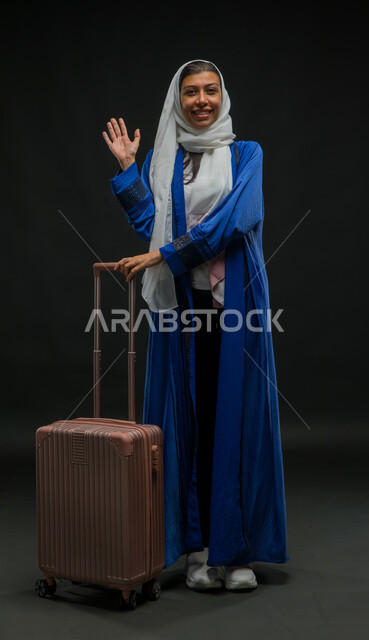 التلويح باليد والتعبير عن الوداع, قضاء وقت ممتع مع الأصدقاء, ايماءات السرور والسعادة بالرحلة السياحية في وقت الإجازة, بورتريه لإمرأة عربية خليجية سعودية مبتسمة ترتدي عباءة زرقاء تمسك بيدها حقيبة السفر وتنظر إلى الكاميرا, صورة لكامل الجسم, خلفية سوداء