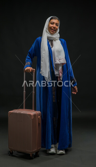 Standing straight and waiting for the plane to arrive, portrait of a Saudi Gulf Arab woman wearing a blue abaya holding a suitcase in her hand, preparing to start the trip during the summer vacation with enthusiasm, the concept of tourism and taking recreational tours during vacation time, full-length body portrait, black background
