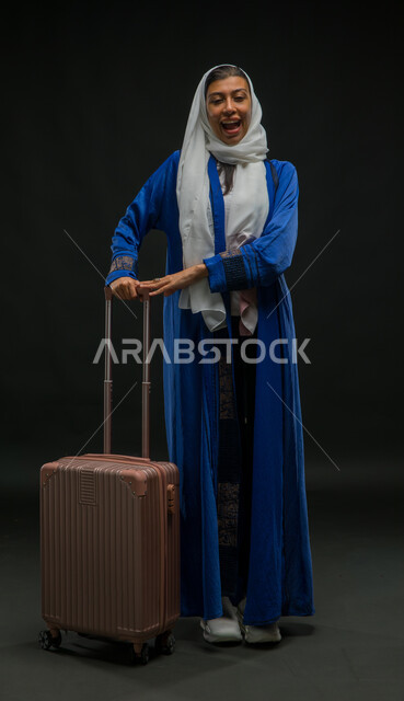 الوقوف باستقامة وانتظار موعد الطائرة, بورتريه لإمرأة عربية خليجية سعودية ترتدي عباءة زرقاء تمسك بيدها حقيبة السفر, الاستعداد لبدء الرحلة في العطلة الصيفيه بحماس، مفهوم السياحة والقيام بالجولات الترفيهية في وقت الإجازة, صورة الجسم بالطول الكامل, خلفية سوداء