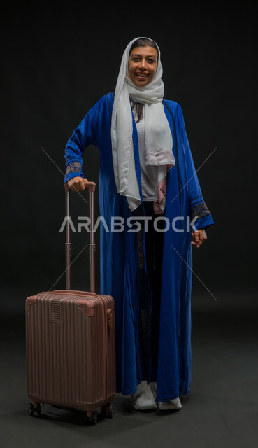 The concept of tourism and taking recreational tours during vacation time, standing straight and waiting for the plane to arrive, portrait of a Saudi Gulf Arab woman wearing a blue abaya holding a travel bag in her hand, preparing to start the trip on the summer vacation with pleasure, full-length body portrait, black background