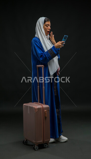 التأخر عن موعد الطائرة بإيماءات الدهشة والانزعاج, مفهوم السياحة والسفر, بورتريه لإمرأة عربية خليجية سعودية ترتدي عباءة زرقاء تمسك الهاتف المحمول, استخدام التقنيات والتطبيقات السياحية, الاستغراب والتوتر لسماع الأخبار السيئة, صورة لكامل الجسم , خلفية سوداء