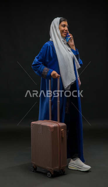 إجراء مكالمة صوتية عبر الهاتف المحمول, التواصل مع الأصدقاء عبر وسائل السوشال ميديا, بورتريه لسيدة عربية خليجية سعودية ترتدي عباءة زرقاء تمسك حقيبة السفر, الاستعداد لبدء الرحلة السياحية بإيماءات البشاشة والسرور, صورة الجسم بالطول الكامل, خلفية سوداء